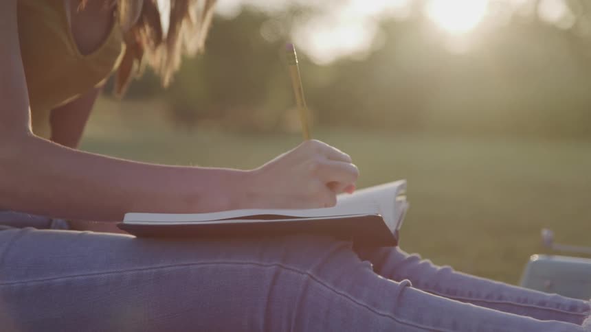 Woman with a yellow shirt and jeans sits on the green grass and writes in a notebook as the sun goes down in the background