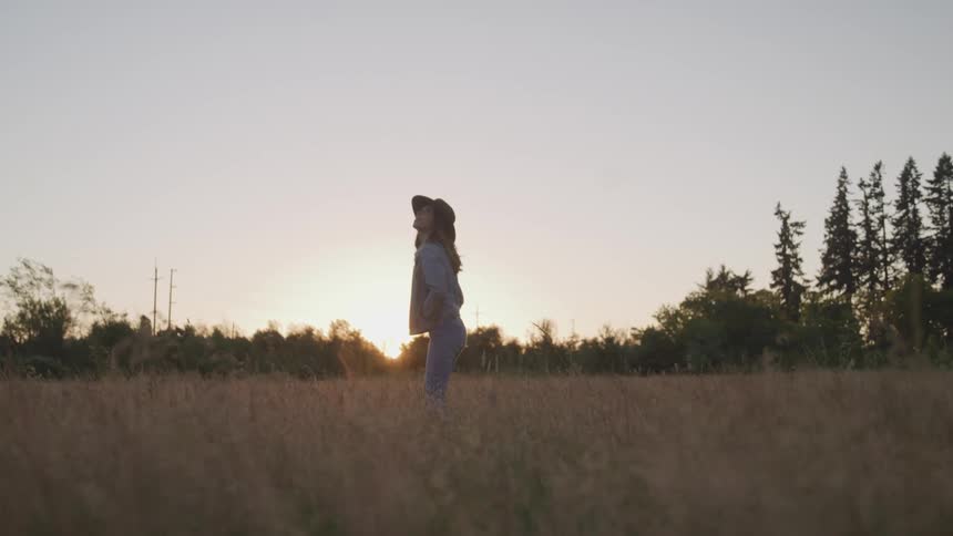 A woman with a hat walking in a grassy field with trees and the sky in the background