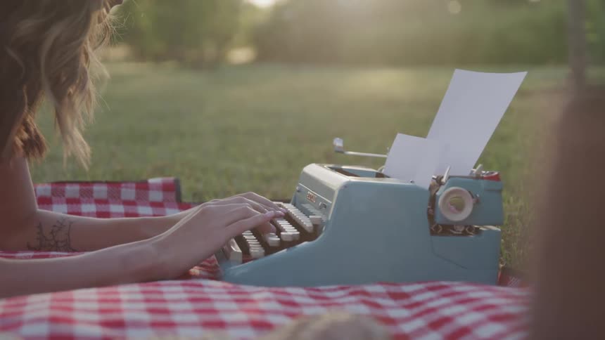 A woman is typing on a typewriter while laying down on a checkered blanket in a grassy lawn