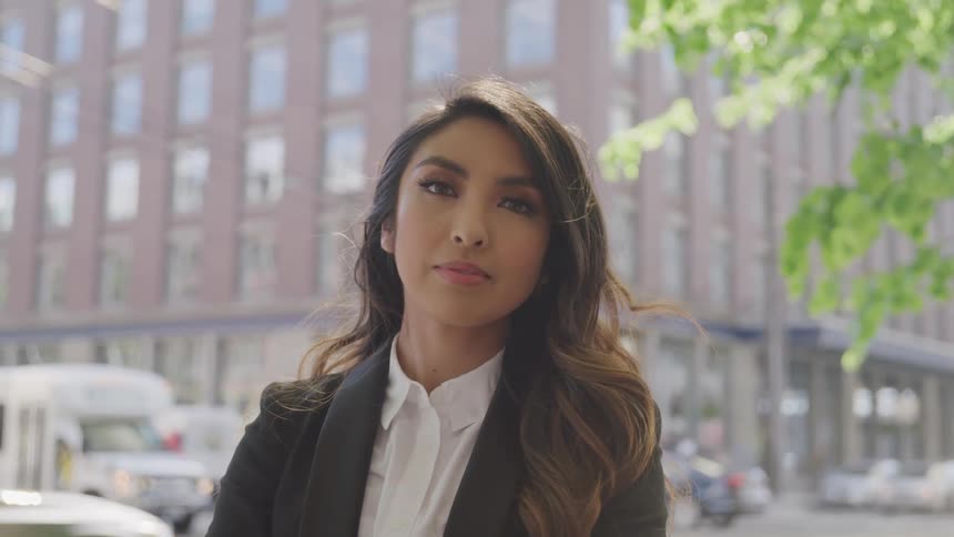 Brunette woman with wavy hair a shirt and a black blazer faces the camera with eye contact in the busy city street with vehicles and buildings in the background