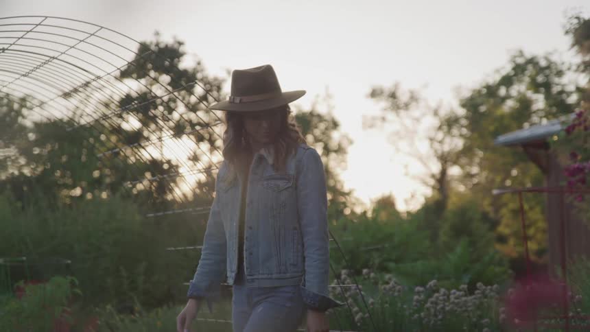 A woman with a hat and jeans jacket walking in a garden with plants and flowers
