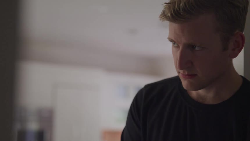 A young man with blonde hair and wearing a black shirt slowly looks up while indoors