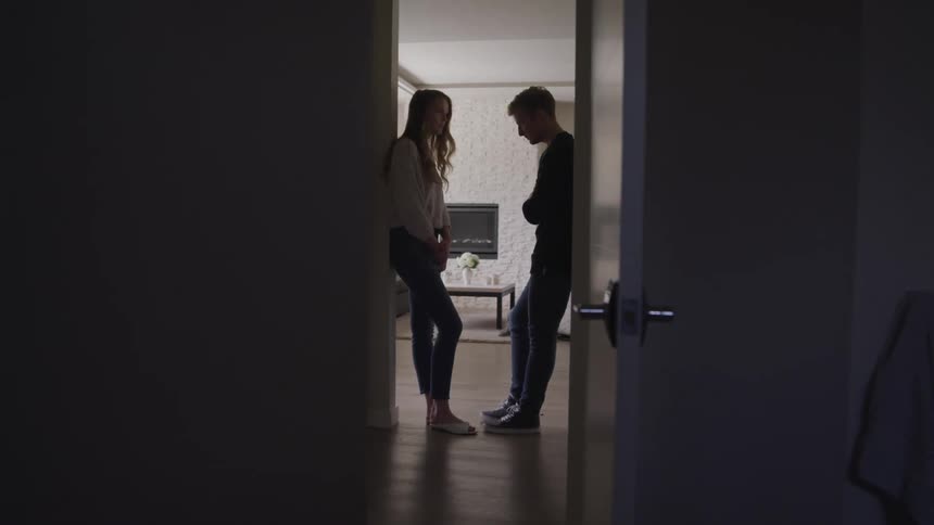 A young woman and man stand facing one another somberly in a doorway