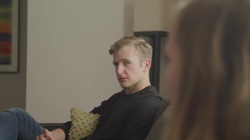 A man and woman sit apart from each other on a couch with a grim look on the mans face