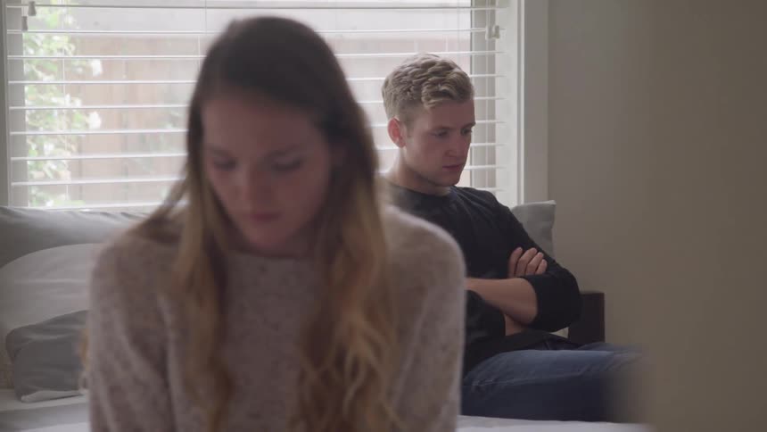 A teenager girl sits with her back to a teenage boy who scowls with his arms crossed