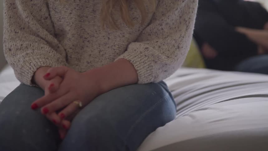 A woman sits on a bed with her hands clasped in her lap behind her a man sits with arms crossed