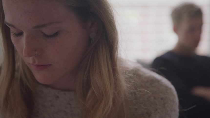 A teenage boy sits on a couch with his arms crossed and a teenage girl with her head down has her back turned away from him