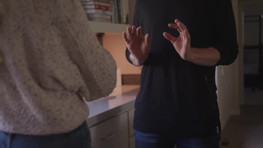 A man and woman stand in a kitchen and talk to one another while using their hands alot