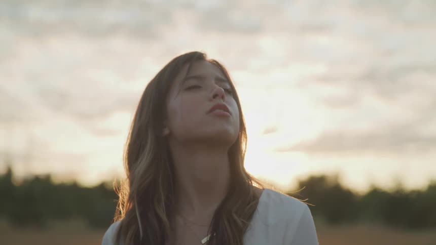 Woman with brown hair and a white shirt looks up with eyes closed as the sun goes down in the background