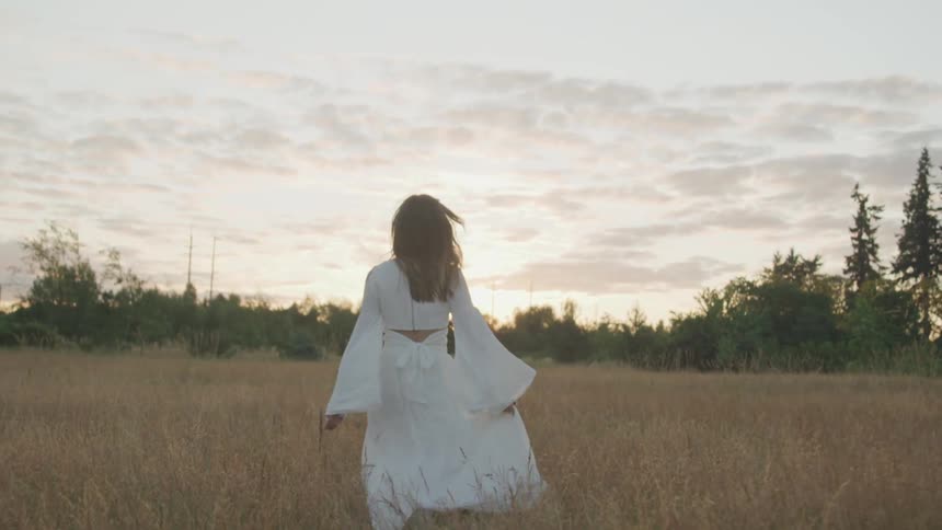 Woman with a white dress runs and jumps happily through dry tall grass in a field at dusk