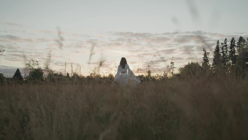 A woman in a white dress walking in a grassy field with trees the sky and clouds in the background