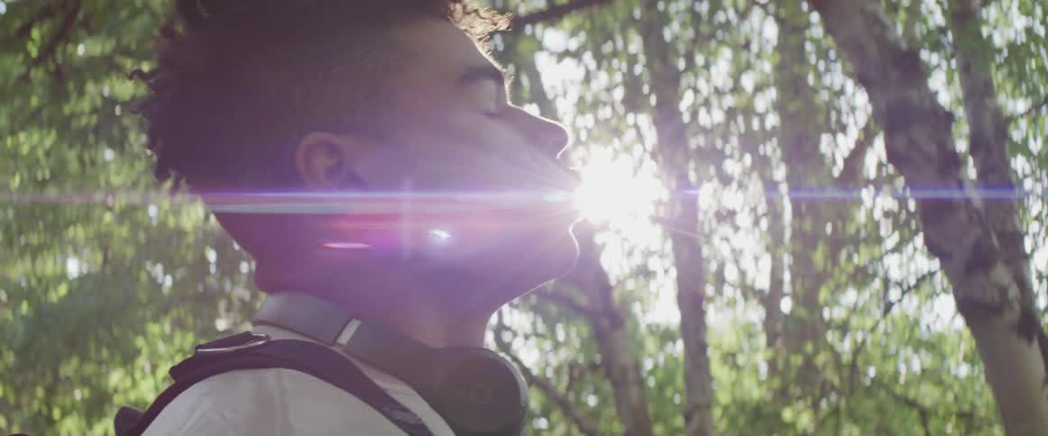 A young man stands outdoors in a woodland with his eyes closed as sunlight shines through the foliage above him
