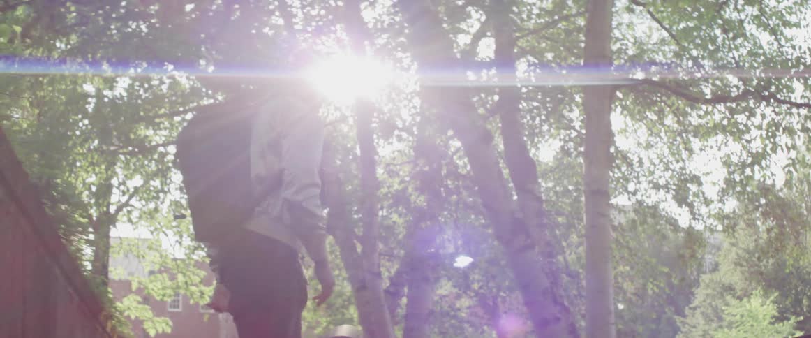 Man wearing backpack dribbles basketball standing near trees sun shines through