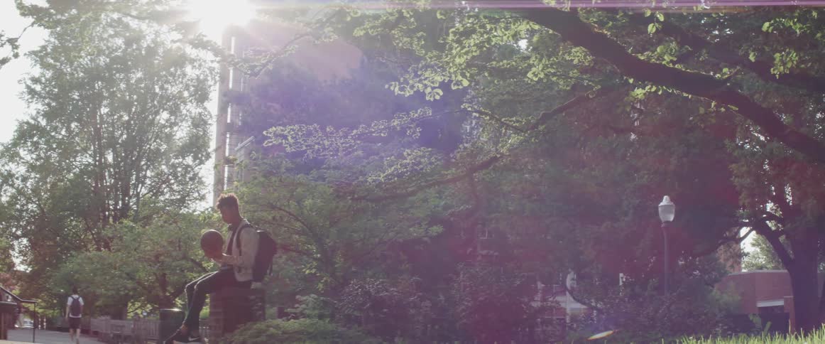Man sits near sidewalk under trees as sun shines past building and through tree leaves