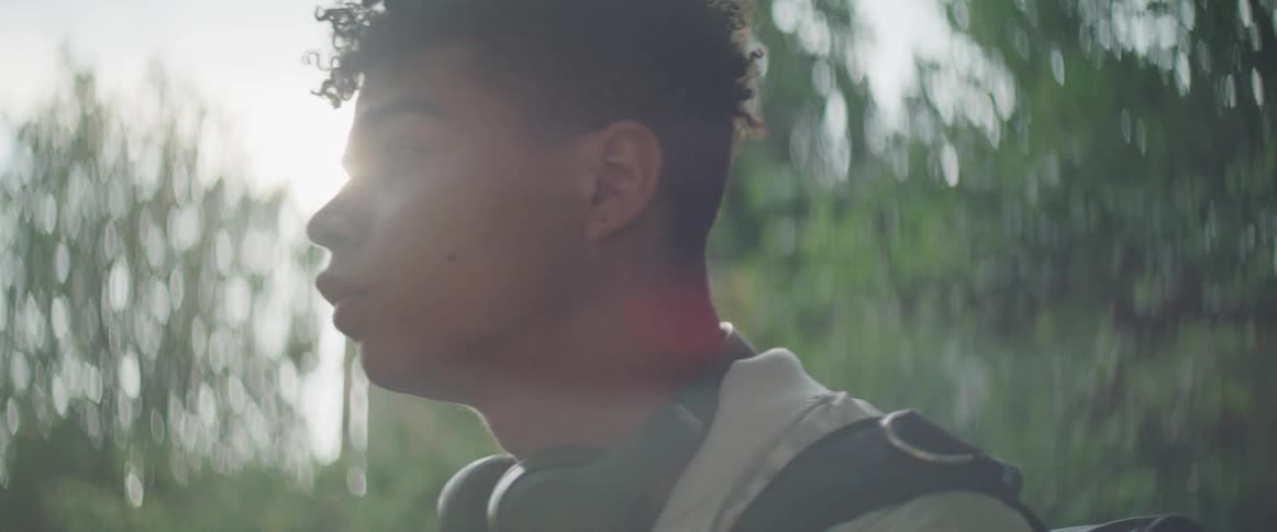 A young black man stands outside wearing a backpack and keeping headphones around his neck