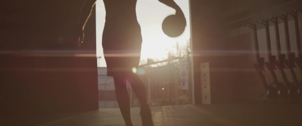 A man dribbles a basketball in a gym while the sun sets through the door