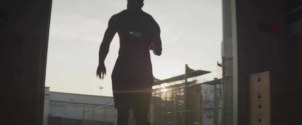 A male dribbles a basketball from inside a large garage or shed to outside of the building
