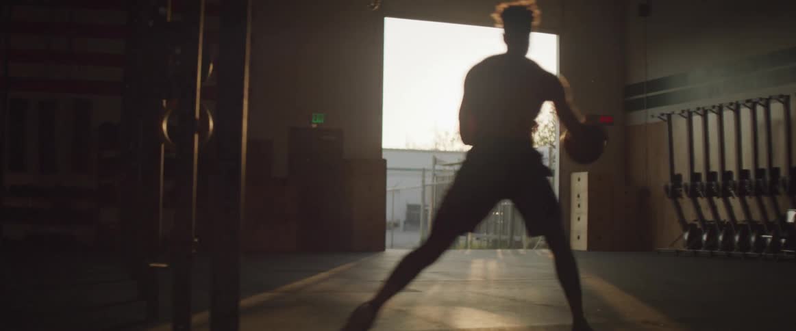 Sunlight shines through the open doorway of a gymnasium onto a shirtless man dribbling a basketball