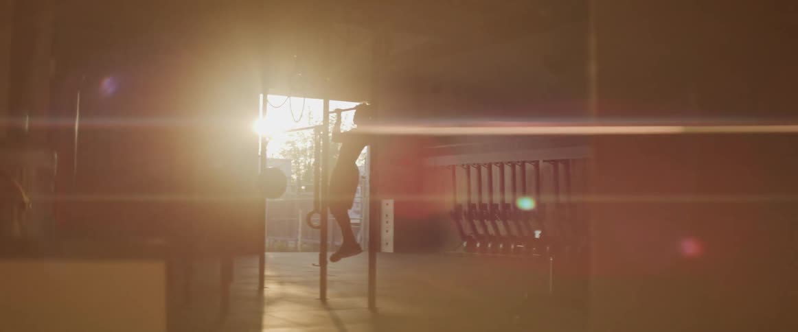 A shirtless man does pullups in a dimly lit gym as sunlight shines in through the open doorway