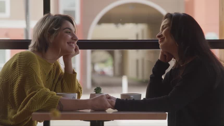 Two women sit at a cafe table holding hands and smiling as they gaze into each others eyes