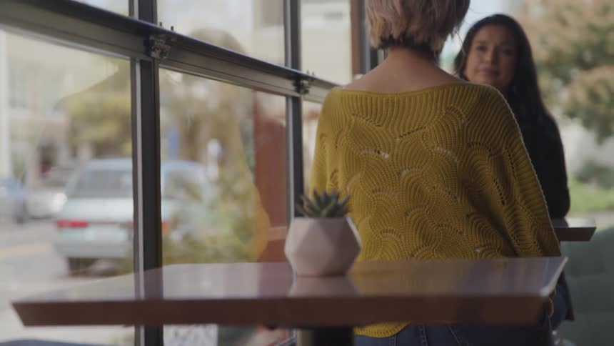 Two women sit together at a cafe table talking next to large windows
