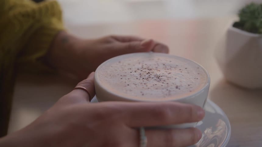 A woman in a yellow blouse sits at her kitchen table and holds a full coffee mug with both hands