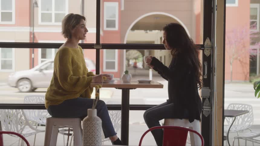 Two women sitting at table drink coffee and talk to each other in a cafe facing the street