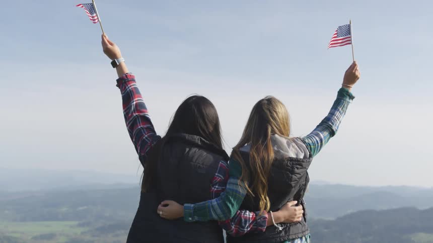 A pair of women standing atop a mountain embracing one another slowly raise small american flags into the air as they look out at the countryside below