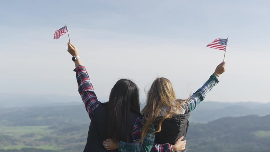 Two women stand together waving american flags on the mountain