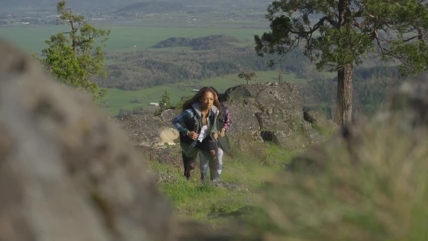 Two women climb grass covered slope with hills and valleys behind them
