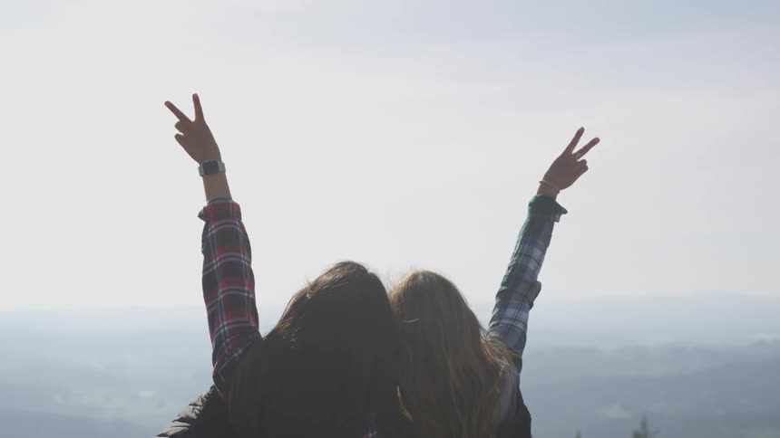 Two women hugging and standing on a cliff with trees the sky and clouds in the background