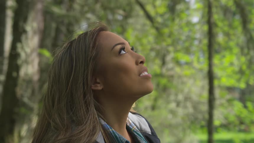 A teenage girl in a forest looks up at the trees and smiles