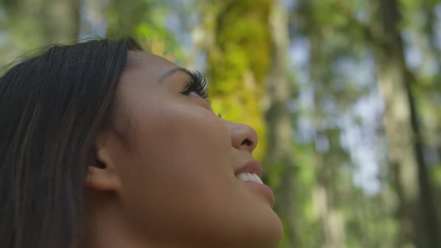 A woman smiles as she looks up at tall trees in a forest