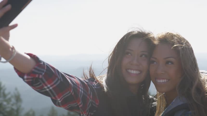 Two women taking a picture with a smart phone with trees the sky and clouds in the background