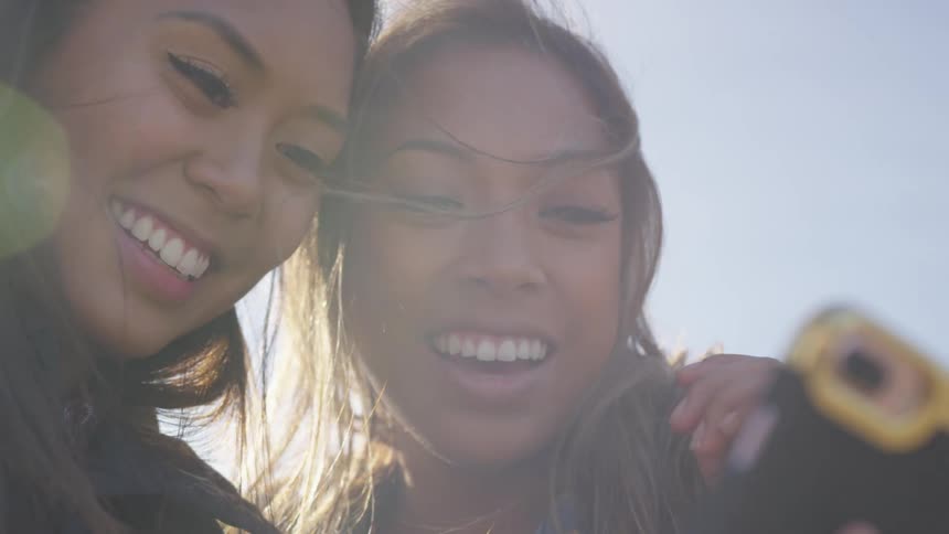 Two friends smile while looking at selfies on a smart phone outdoors