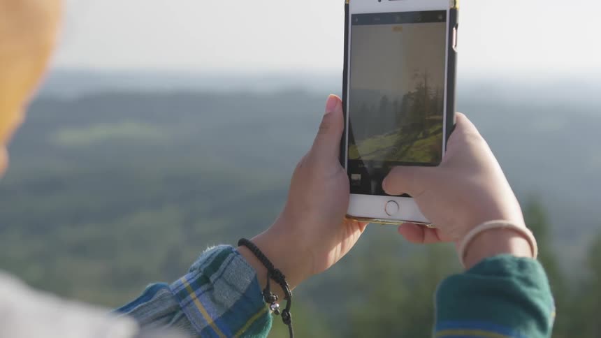 A person with a smart phone taking a picture with trees the sky and clouds in the background
