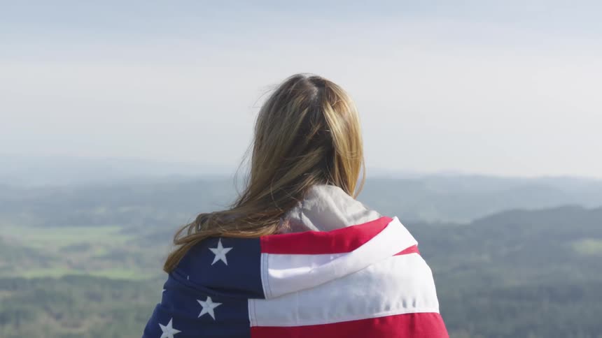 A woman with an american flag wrapped around her shoulders looks out over a forested countryside from high altitude