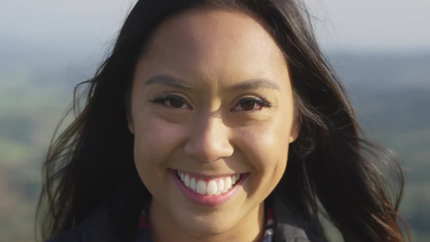 A young woman is smiling and staring into the camera while standing outdoors with a landscape behind her