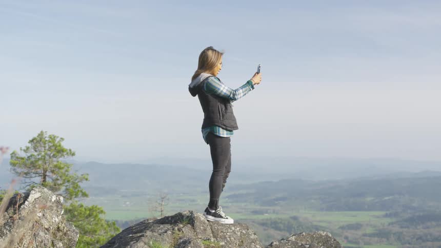 A young woman stands atop a rock that overlooks a large valley as she looks at her phone