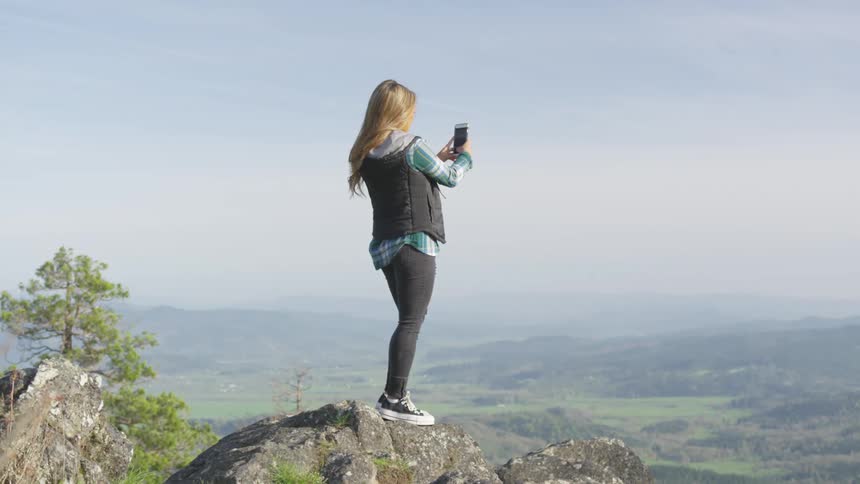 A woman stands on top of a mountain and uses her cell phone to take a photo of the scenery