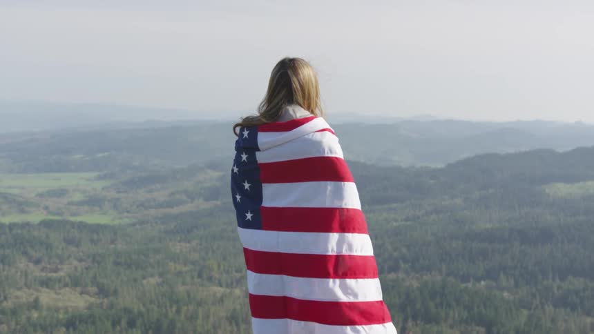 A woman stands at a vantage point above a forest with an american flag around her shoulders