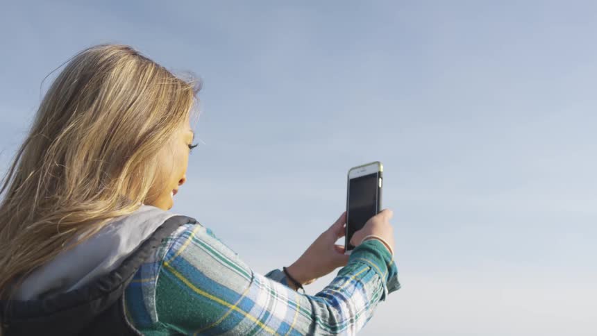A smiling woman standing on a cliff raises her smartphone to take a photograph of the forested countryside below