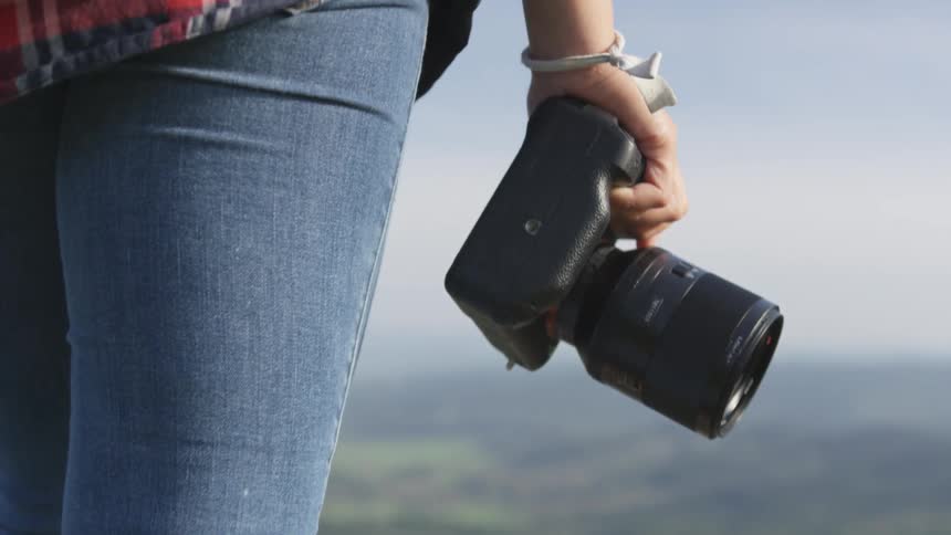 A woman carrying a camera in one hand steps atop a large stone on the the edge of a mountain overlooking a vast countryside