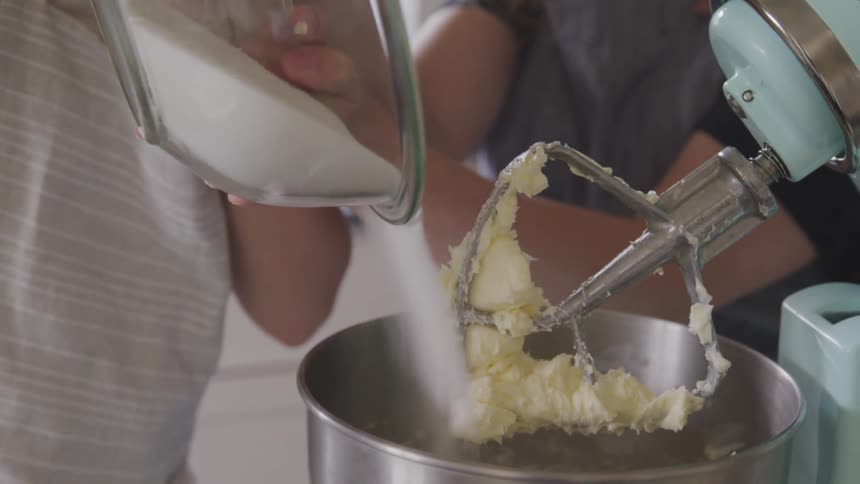 Two people pour flour in the bowl of a mixer in the kitchen