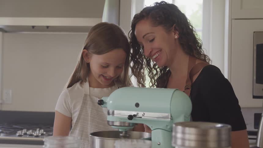 Mother and daughter standing in kitchen smiling and laughing as they watch the beater of the mixer stir the contents of the ingredients in a silver mixing bowl