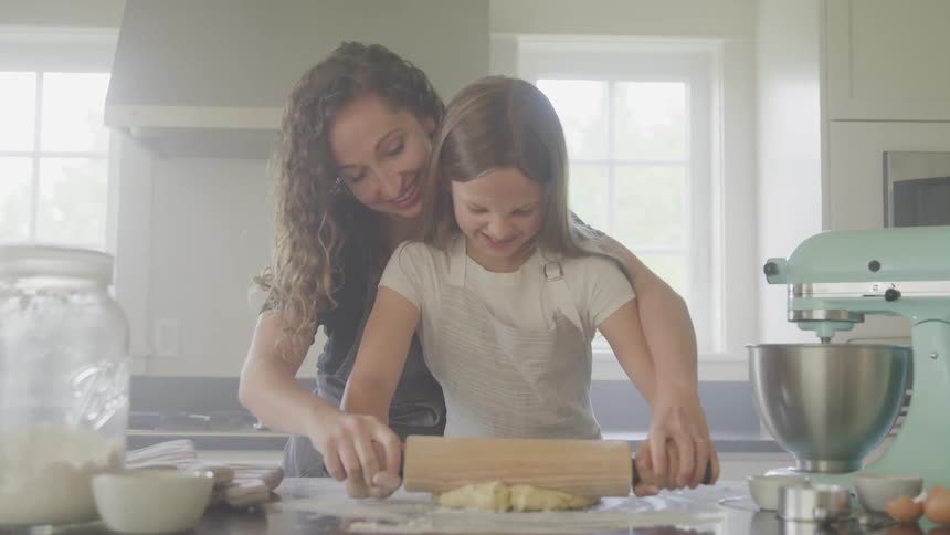Mother helps daughter roll out dough on kitchen counter