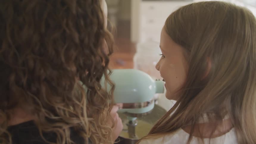 Two girls standing and smiling in a kitchen using a mixer