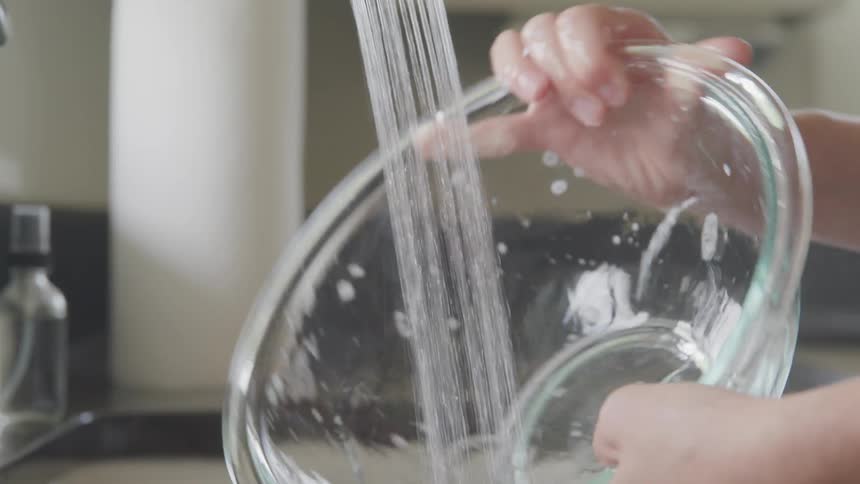 A person rinses the soap off of a glass bowl using a faucet