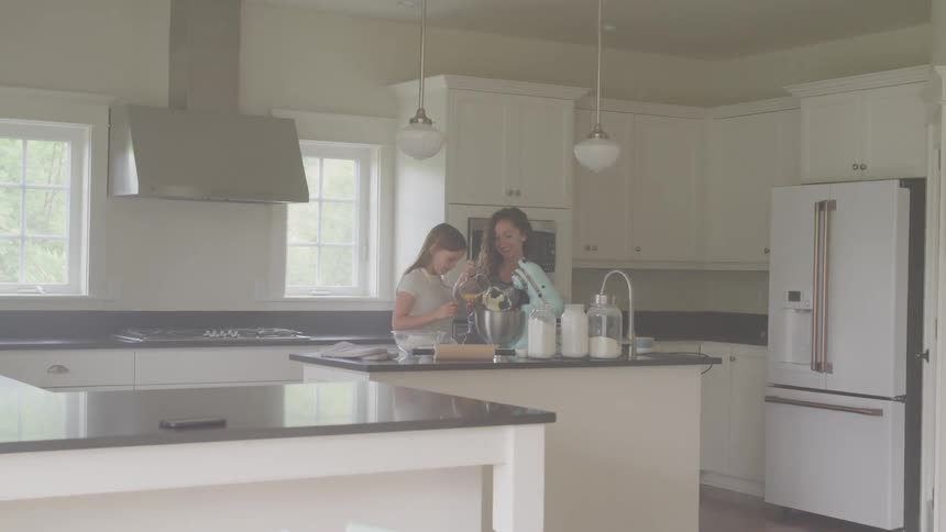 Two girls in a kitchen next to a counter with a refrigerator with windows in the background