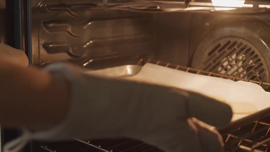 Female wearing oven mitts places a cookie sheet with sugar cookie dough on top of parchment paper into a lit oven for baking